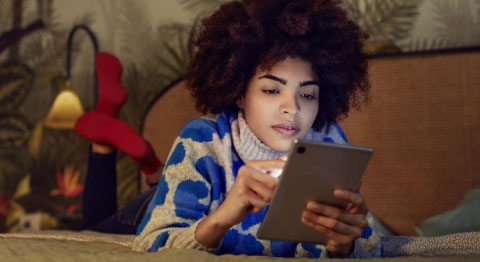 Lockige Frau liegt auf dem Bauch auf ihrem Bett und schaut in ihr Tablet
