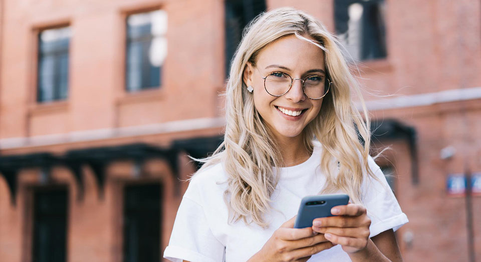 Frau mit blonden langen Haaren und Brille hält Handy und lächelt