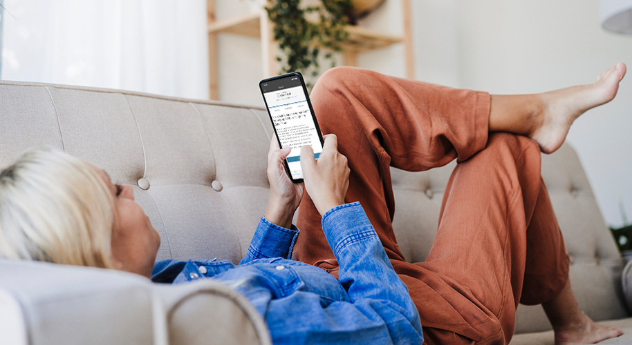 Blonde Frau  in blauer Bluse sitzt mit dem Smartphone zufrieden lächelnd in einem dunklen Wohnraum