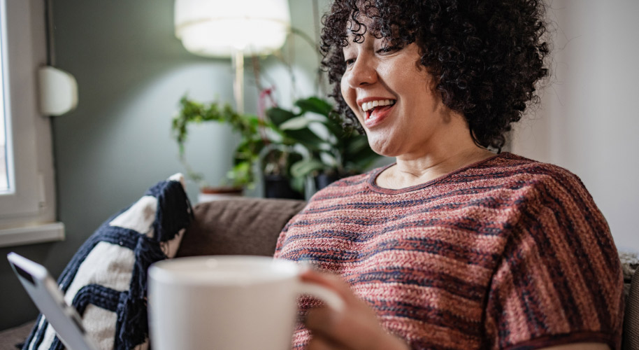 Frau mit Locken sitzt lachend vor dem Laptop auf dem Sofa