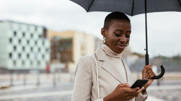 Frau im Mantel mit Regenschirm und Smartphone in der Hand